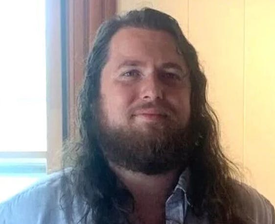 A person with long hair and a beard, wearing a light-colored shirt, standing indoors near a window.