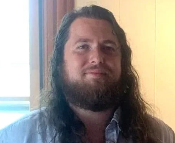 A person with long hair and a beard, wearing a light-colored shirt, standing indoors near a window.