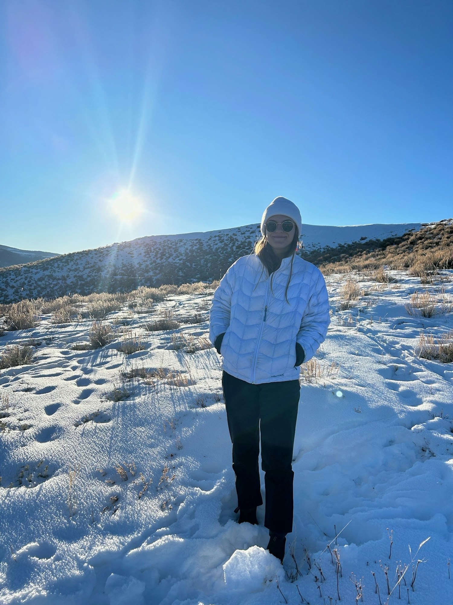 A person in a white jacket and sunglasses stands on snow-covered ground with a sunlit background and clear blue sky.