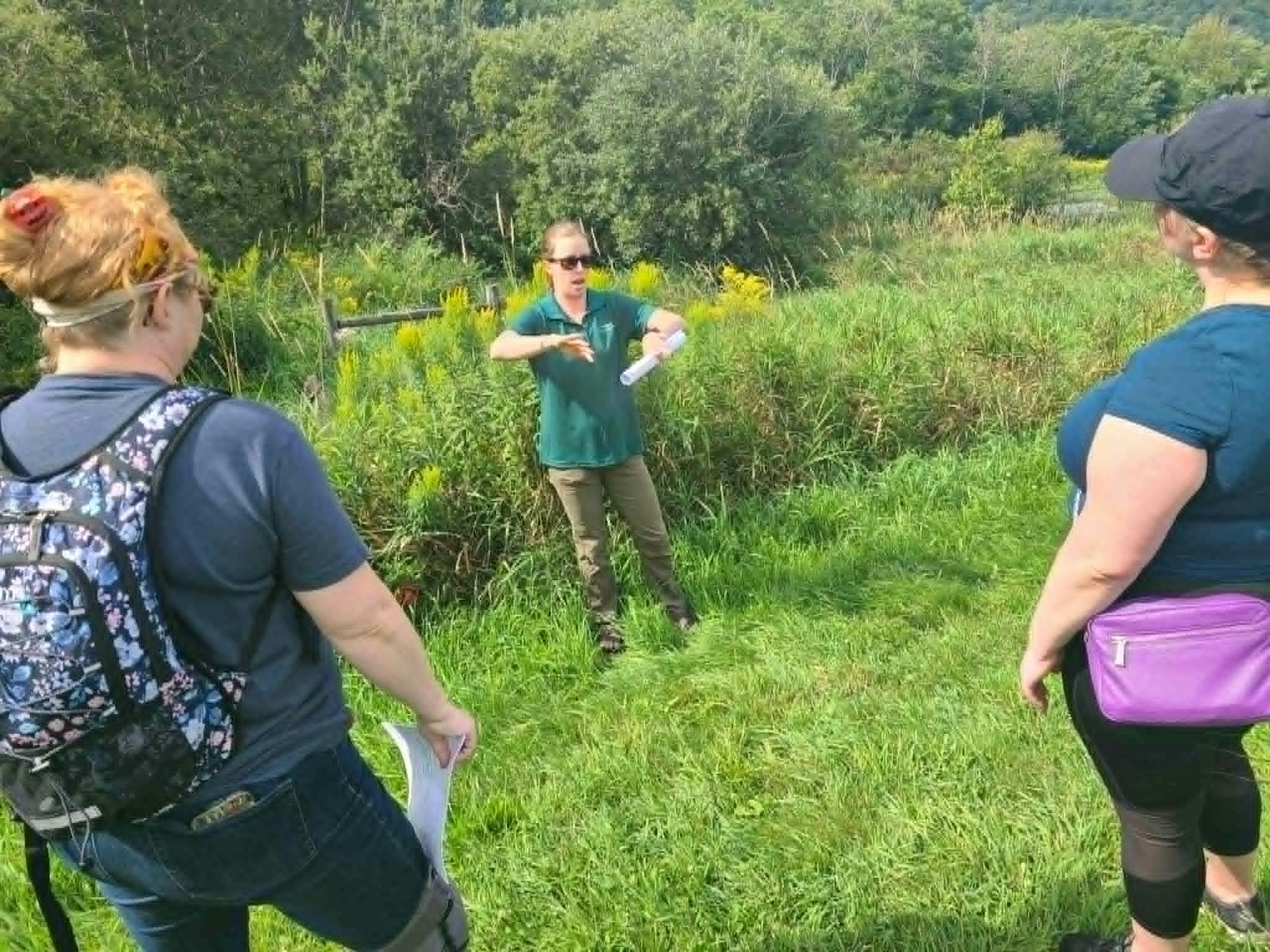 A person is giving an outdoor presentation or lesson to two others amidst greenery.