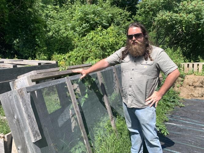 A person with long hair and a beard wearing sunglasses stands next to wooden structures in a green, outdoor setting.