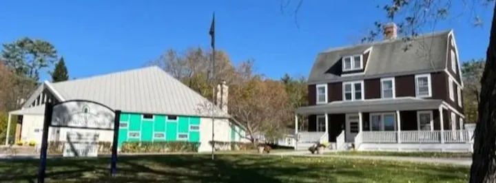 The image shows two buildings: a modern structure with a green facade and a traditional house with a porch, set against a blue sky.