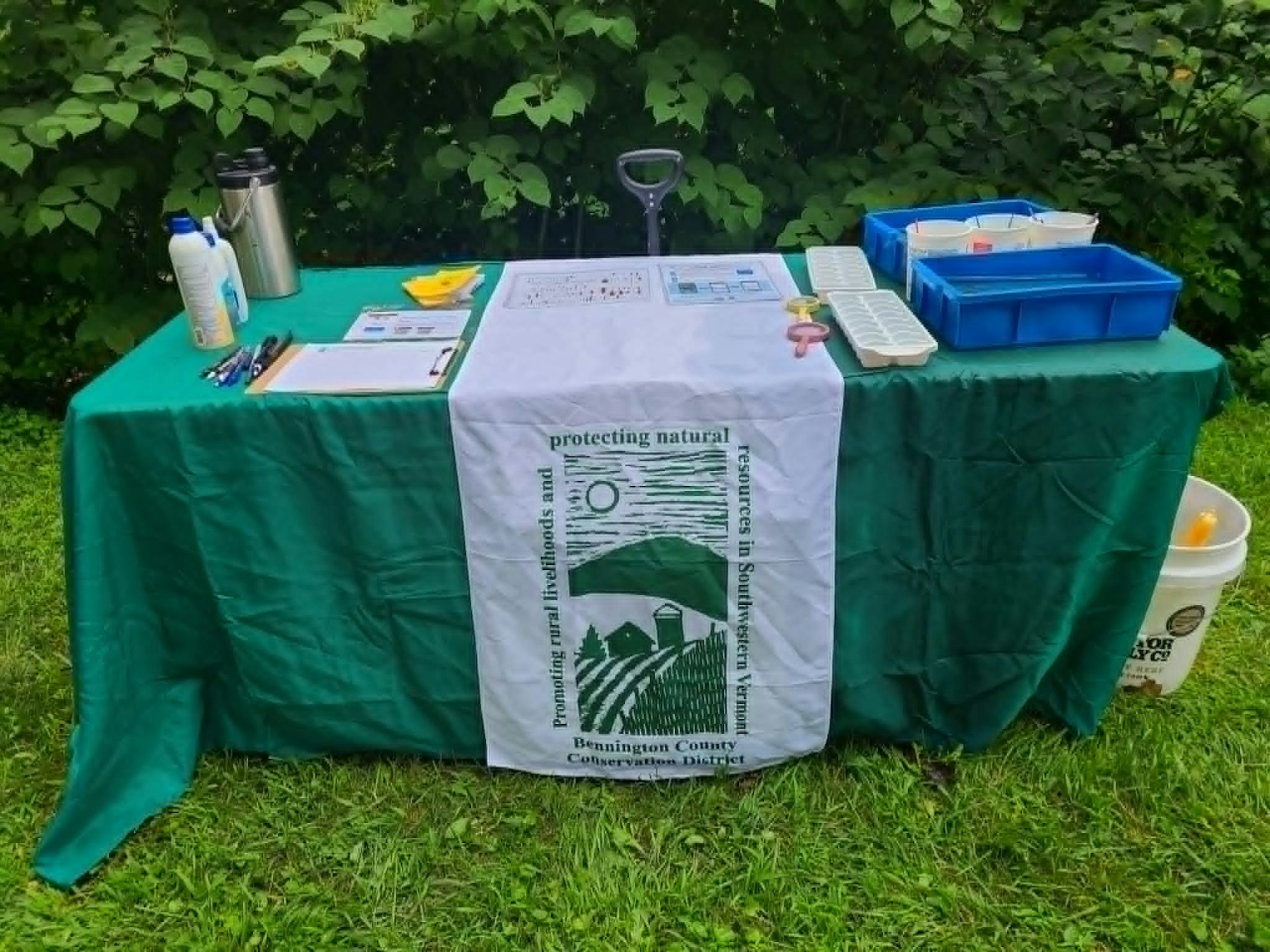 A green table with a banner for a conservation district, tools, clipboards, and containers set in a grassy area.