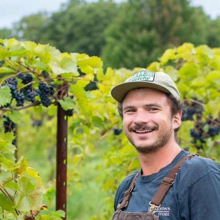 A person in a vineyard wearing a cap and overalls, smiling with grapevines in the background.