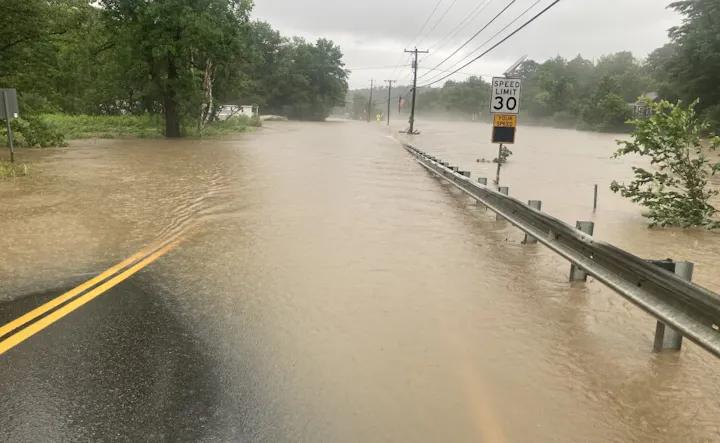 A flooded road with brown water covering the pavement, trees nearby, and a speed limit sign partially visible.