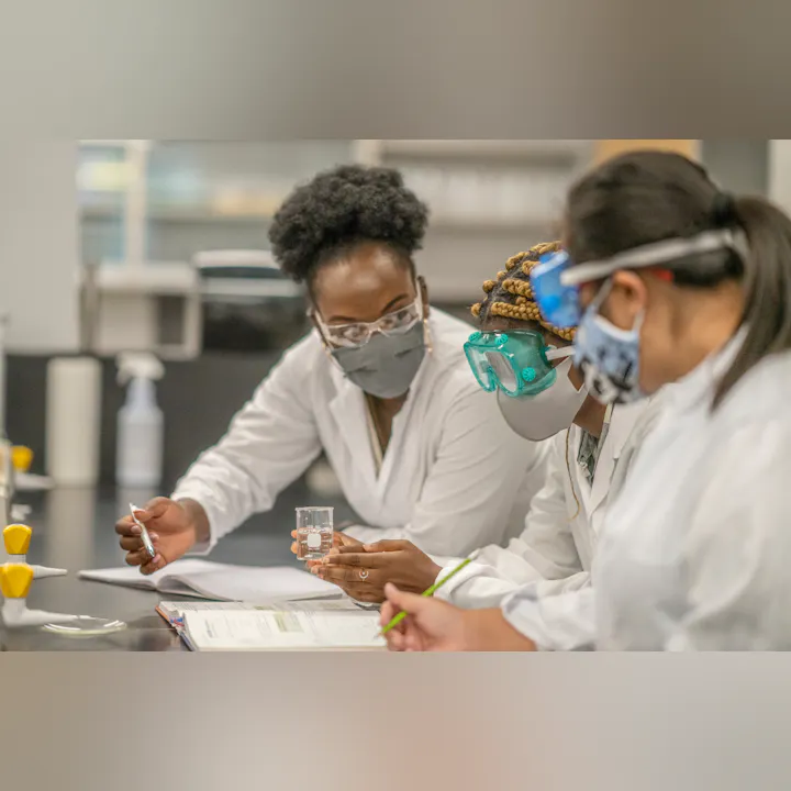 Three individuals in lab coats and safety gear collaborate in a laboratory, analyzing data and holding a beaker.