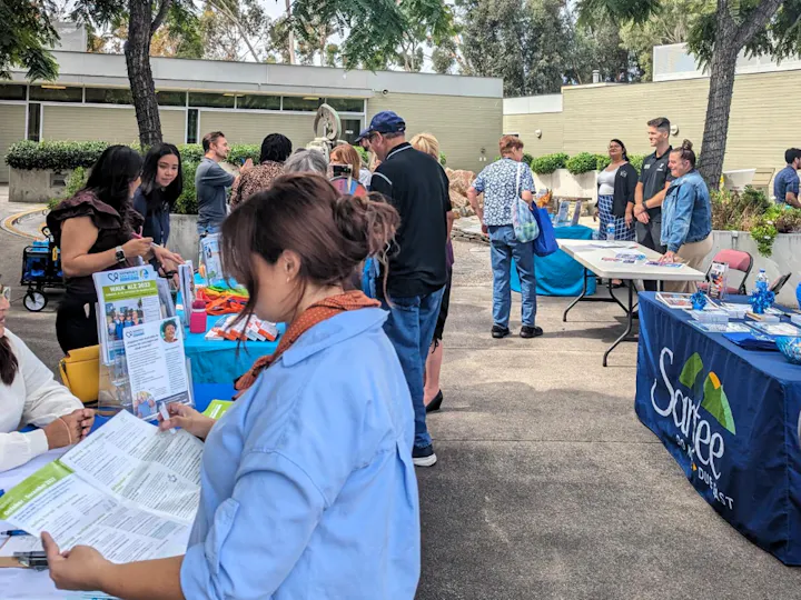 The image shows a community event with tables, people interacting, and informational materials displayed.