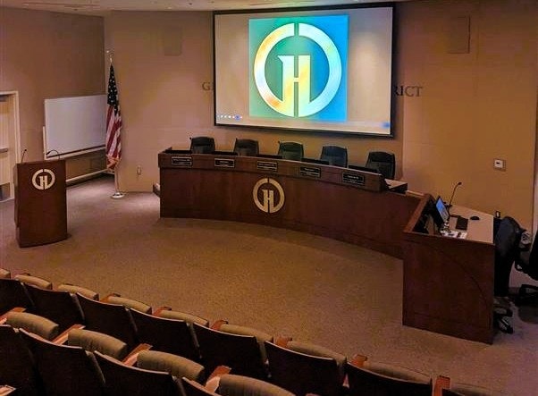 The image shows a meeting room with a large screen, seating, a podium, and a U.S. flag, likely for official meetings.