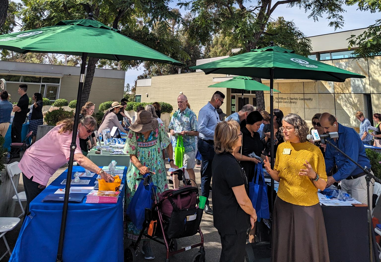 A lively outdoor event with attendees engaging at booths, shaded by green umbrellas, offering resources and information.