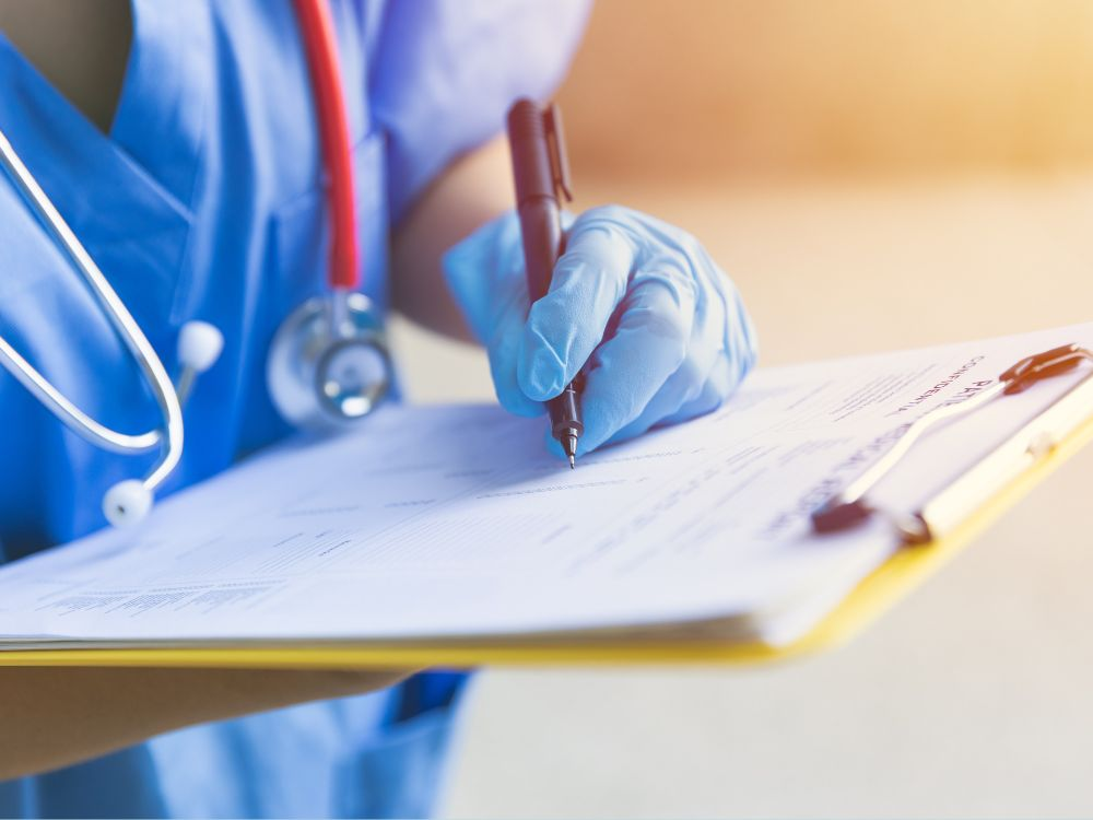 A healthcare professional wearing scrubs and gloves writes on a clipboard with a stethoscope nearby.