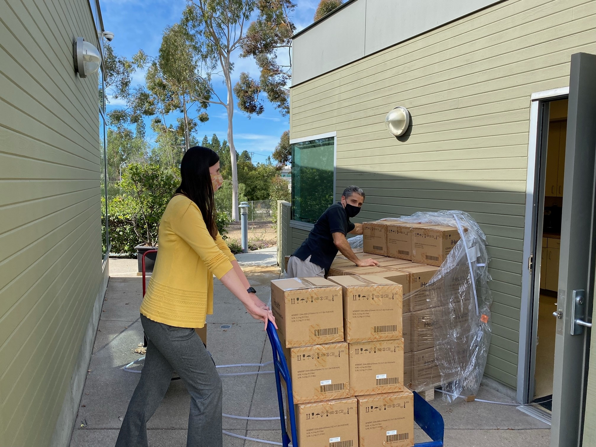 A woman in a yellow sweater and a man in a black shirt are moving boxes on a cart outside a building.