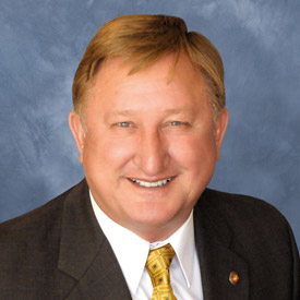 The image shows a smiling man in a suit with a light shirt and patterned tie, against a blue background.