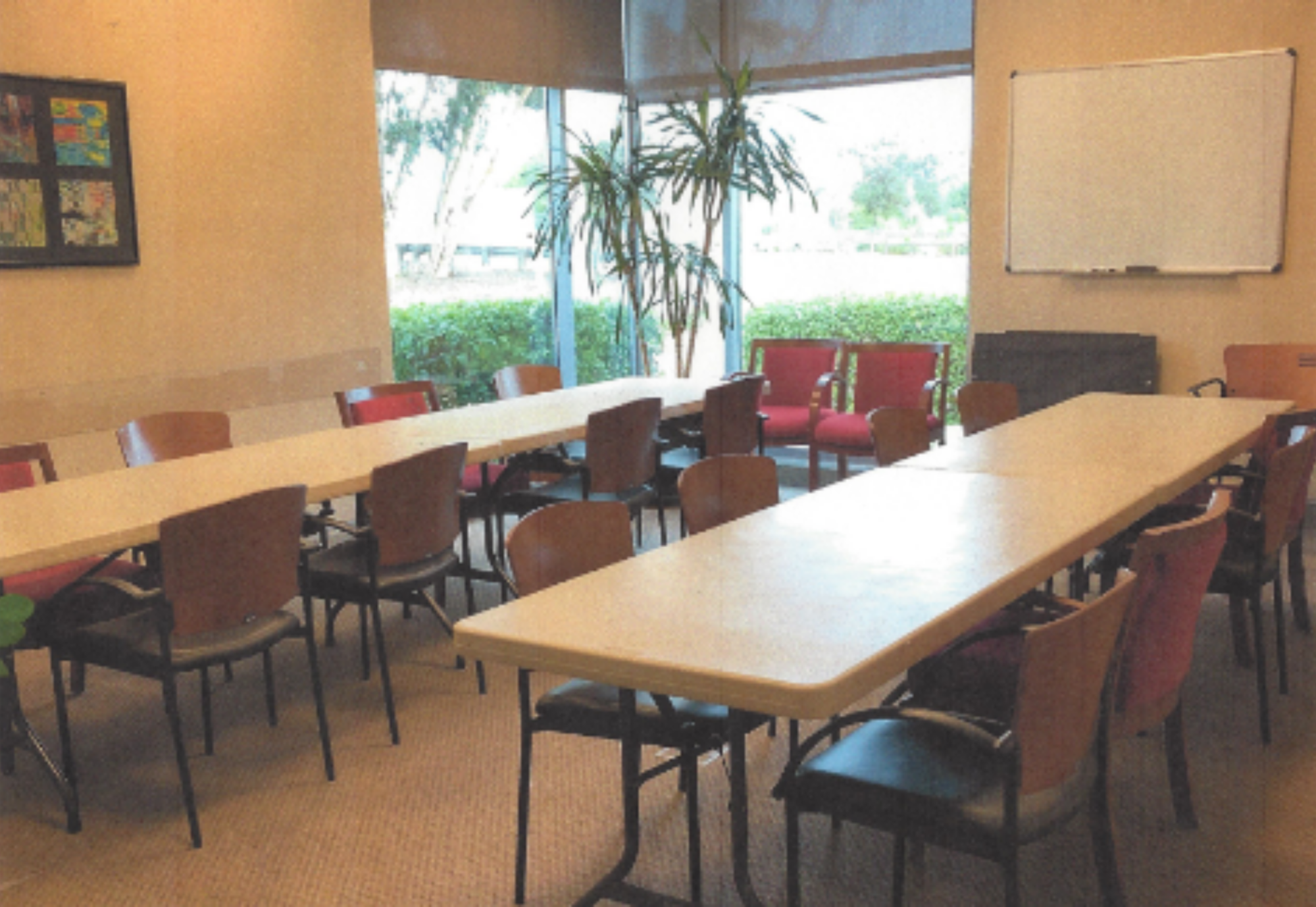 A meeting room with long tables, chairs, a whiteboard, and a large window overlooking greenery.