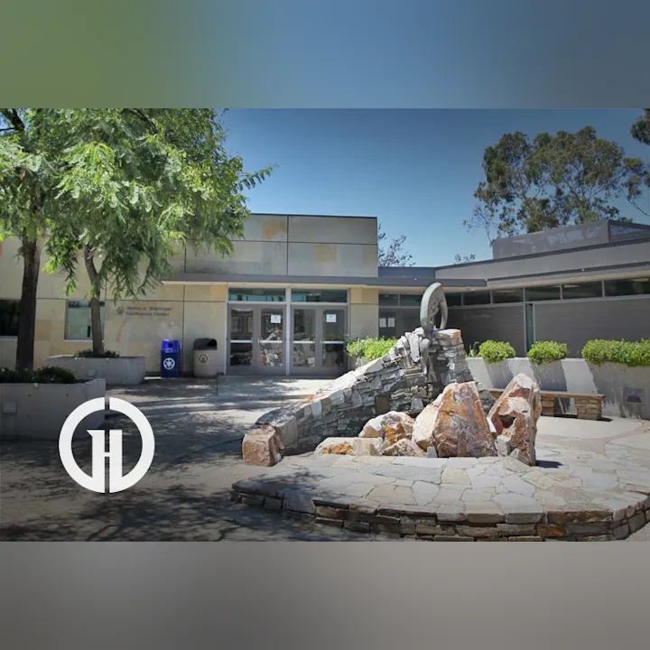 The image shows a modern building entrance with a stone sculpture in front, surrounded by greenery and paved pathways.