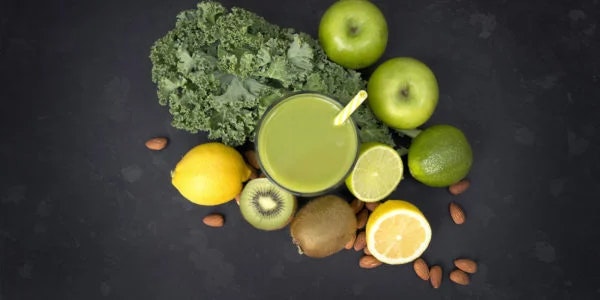 A vibrant green smoothie surrounded by kale, green apples, lemons, kiwi, limes, and almonds on a dark background.