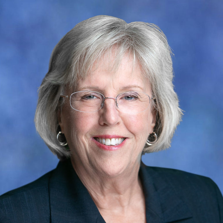 The image features a smiling woman with short gray hair, wearing glasses and a dark blazer, against a blue background.