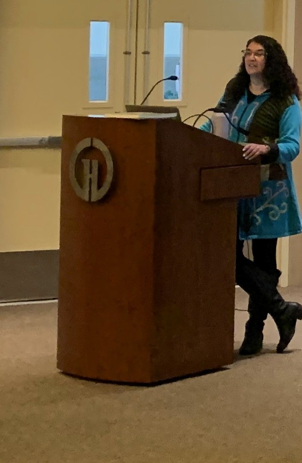 A person stands at a podium, speaking or presenting in a room with large windows and a circular logo in the background.