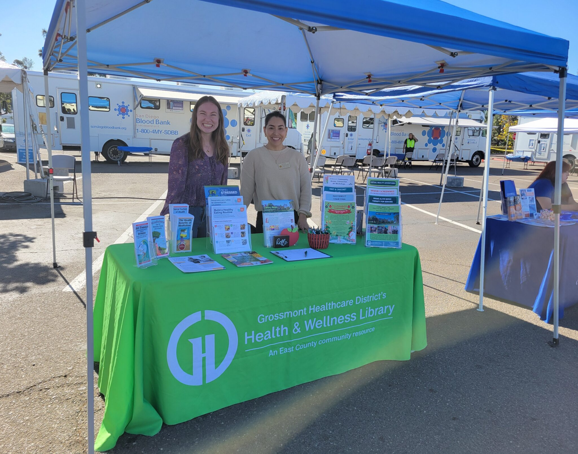 Two women at a booth for Grossmont Healthcare District's Health & Wellness Library at an outdoor event, with informational materials displayed.