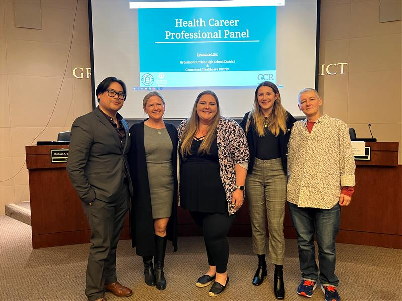 A group of professionals poses at a Health Career Professional Panel event, with a presentation screen in the background.