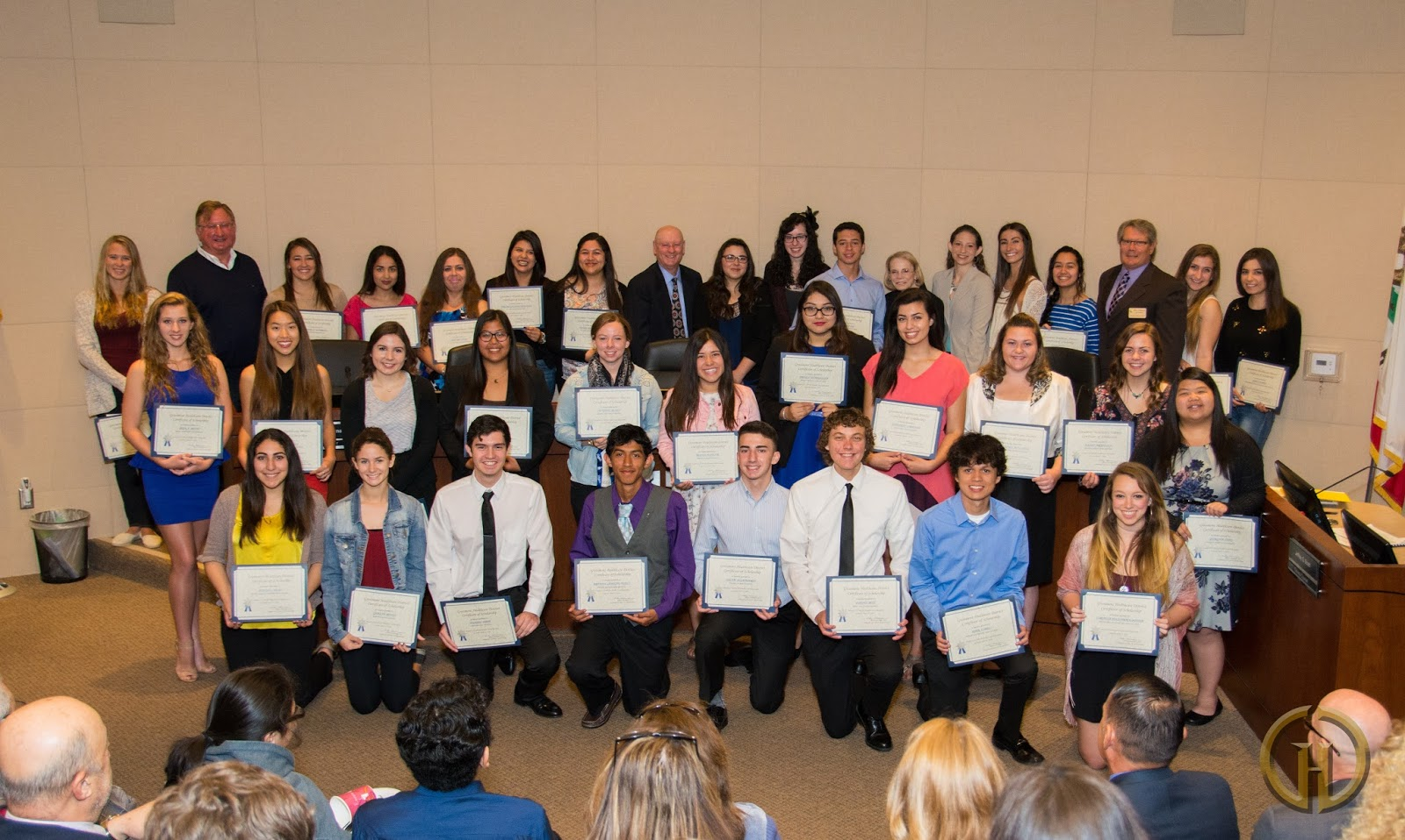 A group photo of students holding certificates, celebrating an achievement, with attendees in a formal setting.