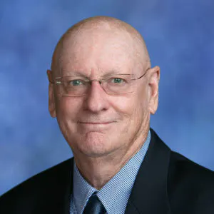 The image shows a man with a bald head, wearing glasses and a suit, against a blue background.