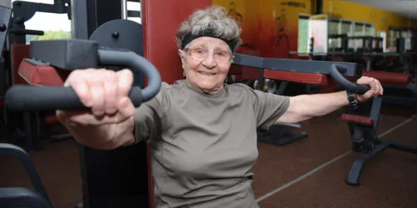An elderly woman exercises on a gym machine, smiling and showcasing her fitness routine in a lively gym setting.