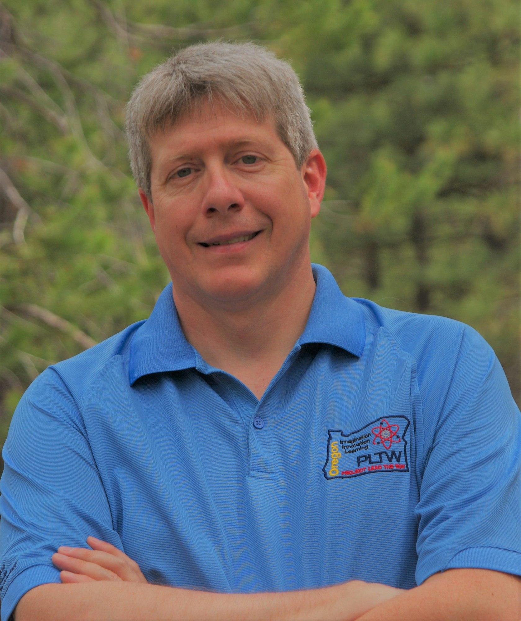 A person in a blue polo shirt with a logo, standing outdoors with trees in the background.