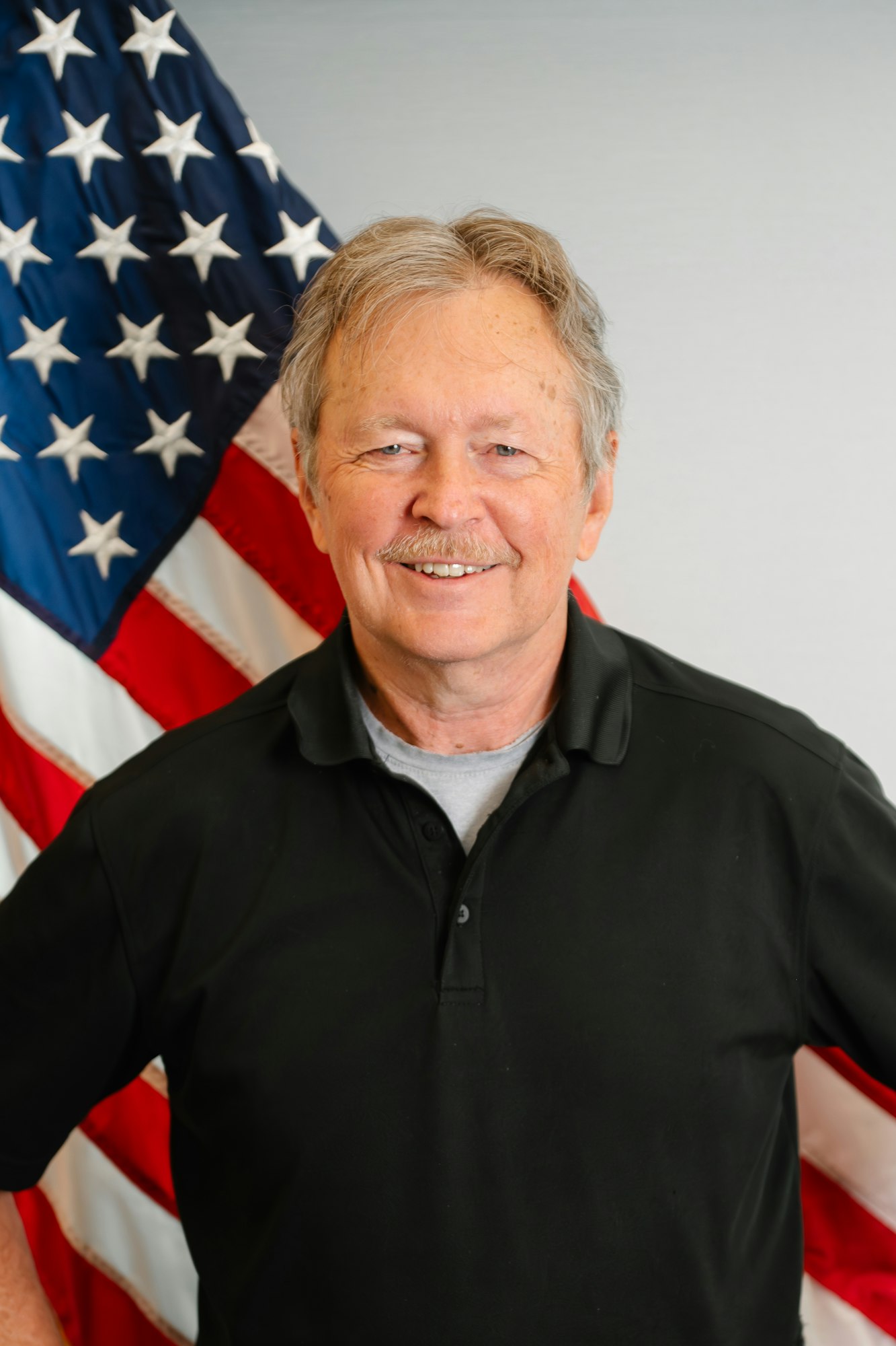 A smiling man stands in front of an American flag, wearing a black polo shirt.