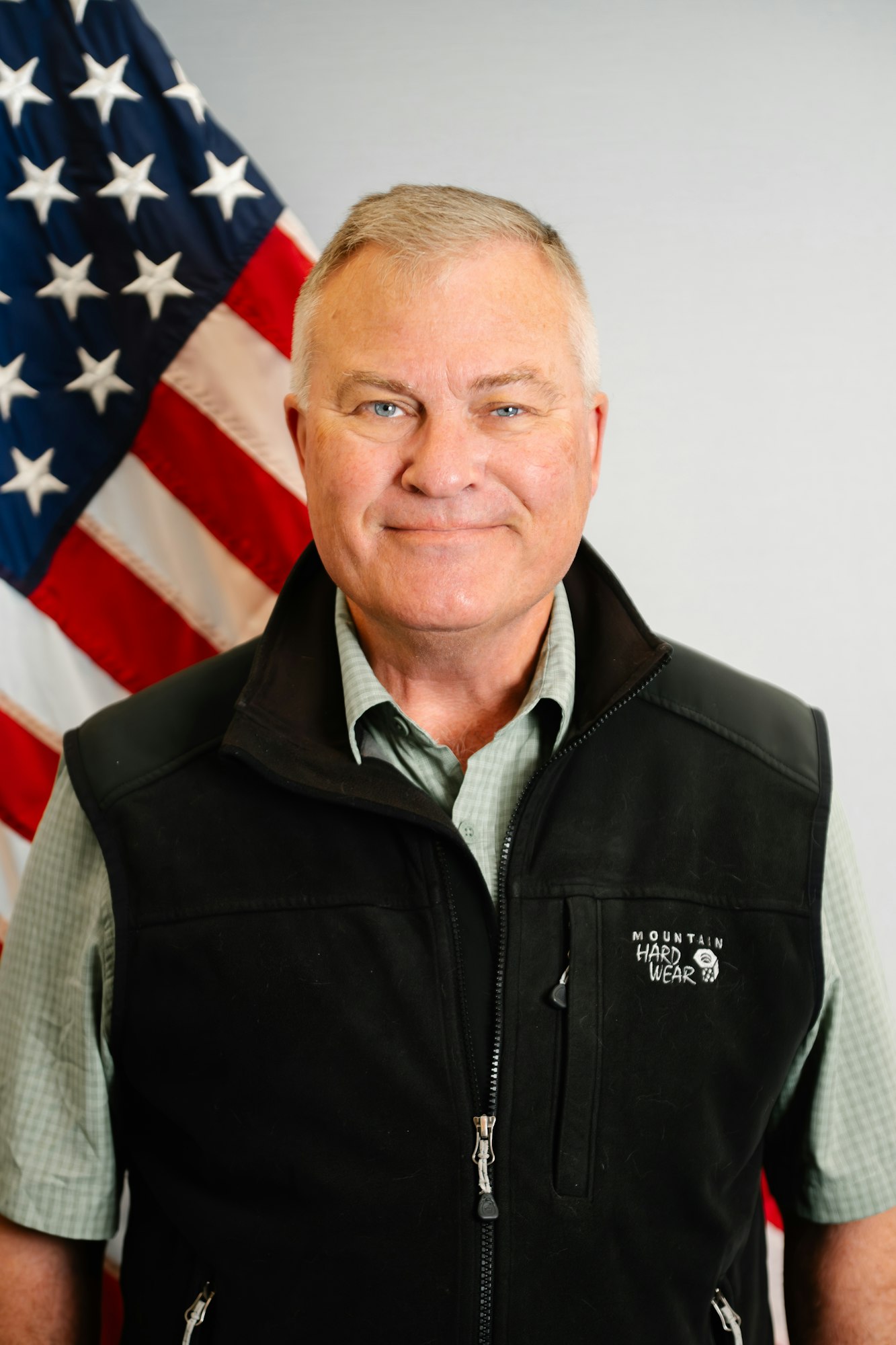A smiling individual wearing a black vest stands in front of an American flag backdrop.