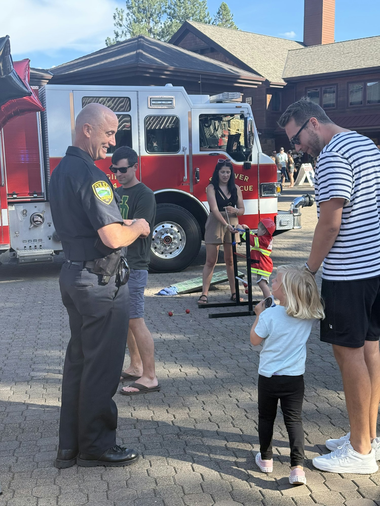 People interacting with a police officer near a fire truck. A child is looking on while adults are engaging in conversation.