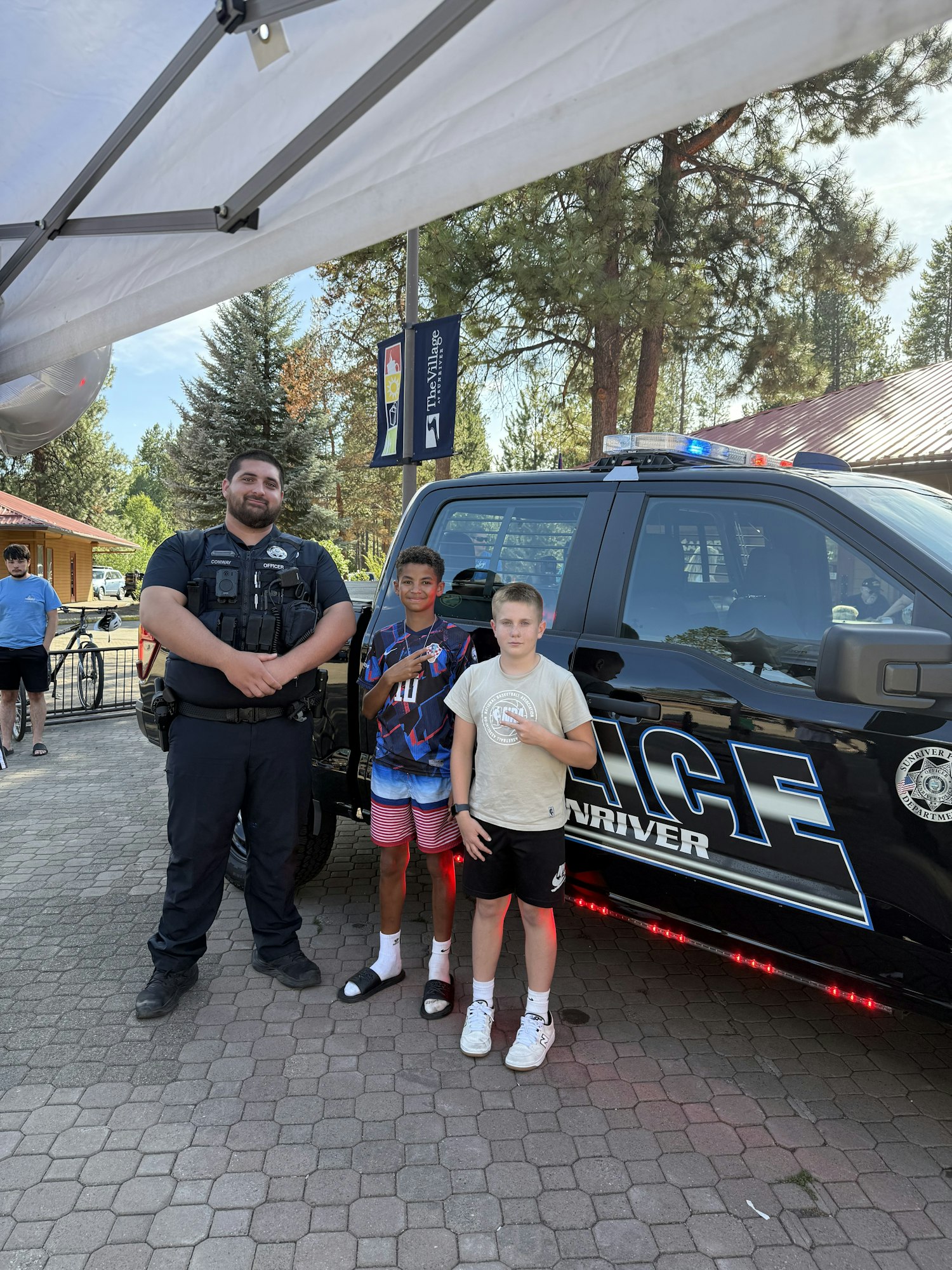 Three people standing in front of a police vehicle, with trees and a banner in the background.
