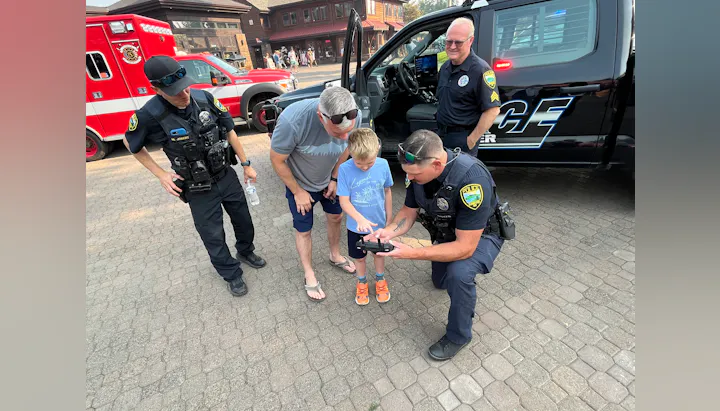 A child interacts with police officers, showing interest in a device near an emergency vehicle while a man supervises.