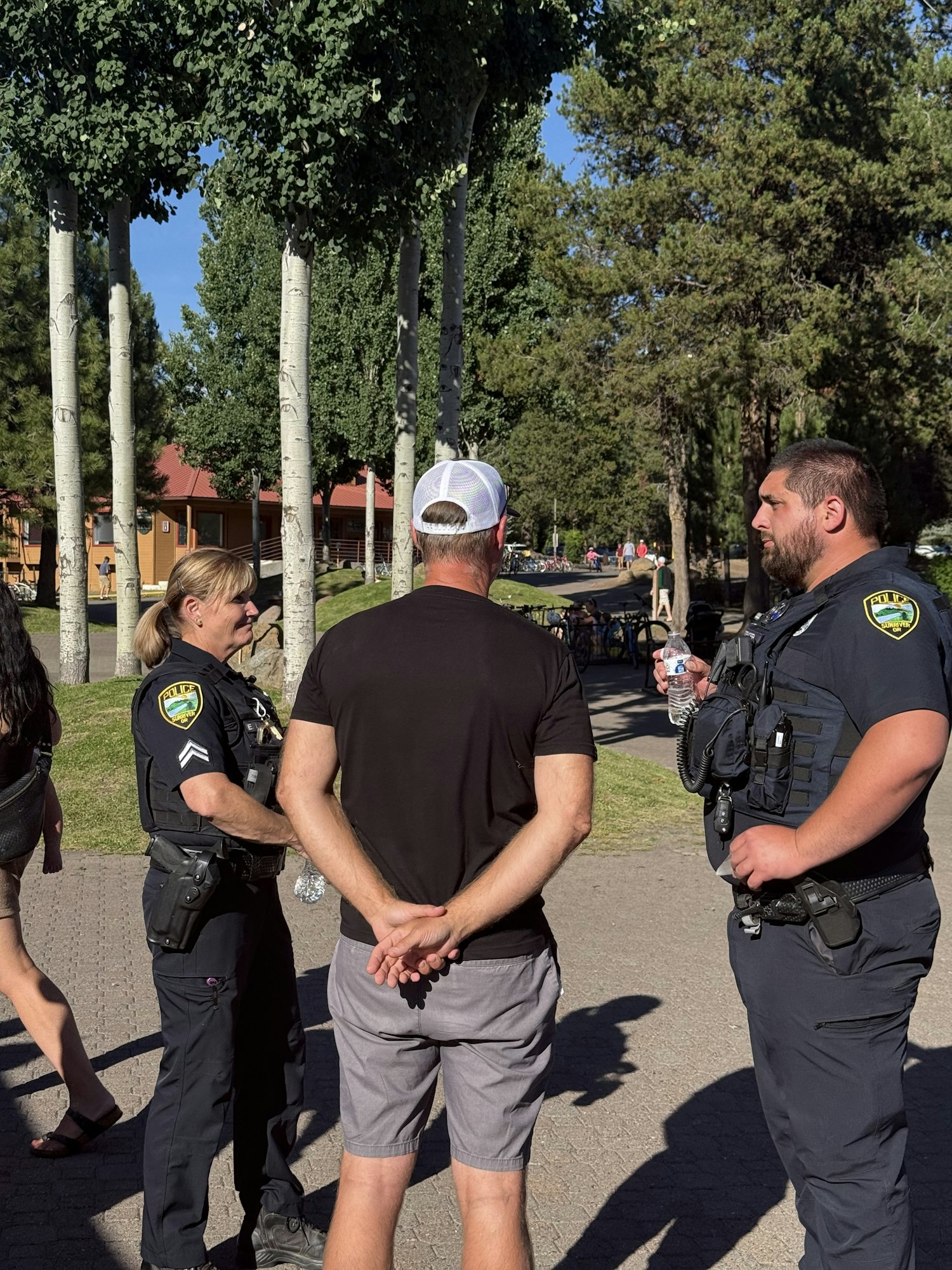 Two police officers talking to a man with hands behind his back in a park setting.