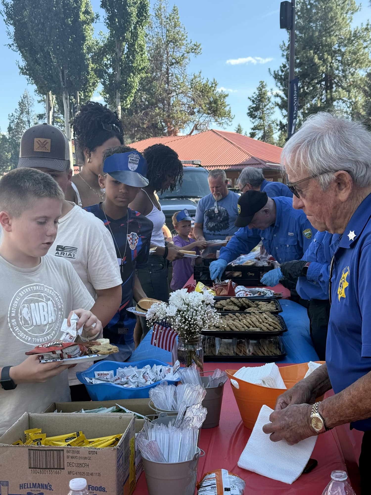 People at an outdoor food table, serving and receiving food, with utensils, snacks, flowers, and an American flag decoration.