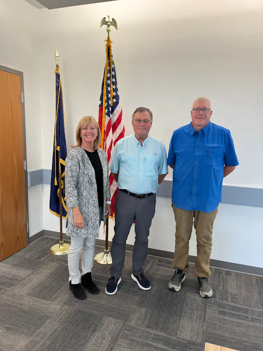 Three people are posing in front of flags, smiling. They appear to be in a formal setting, possibly an office or government building.