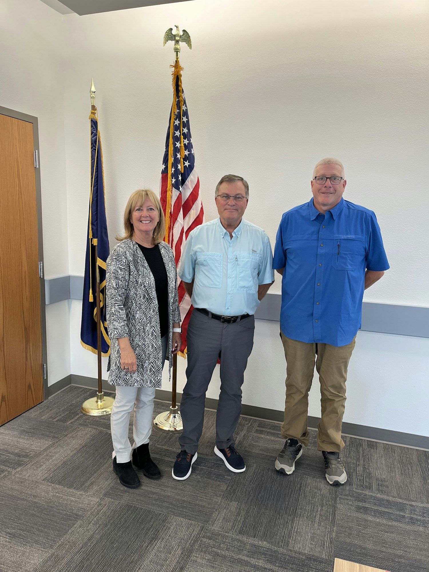 Three people are posing in front of flags, smiling. They appear to be in a formal setting, possibly an office or government building.