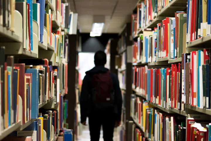 A person stands between rows of colorful books in a library, illuminated by soft overhead lighting.