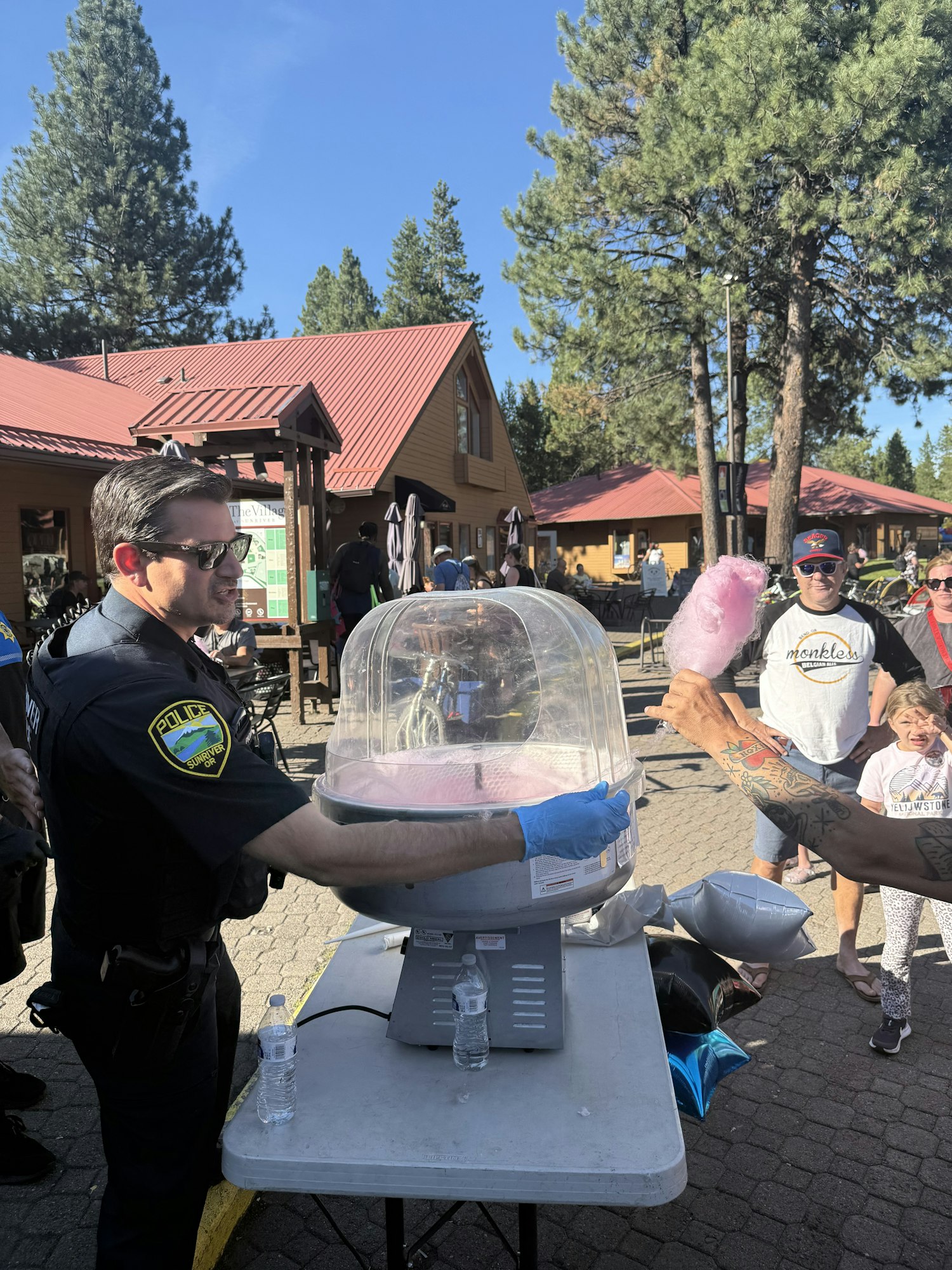 A police officer making cotton candy outdoors, with a crowd watching. There are trees and a building in the background.
