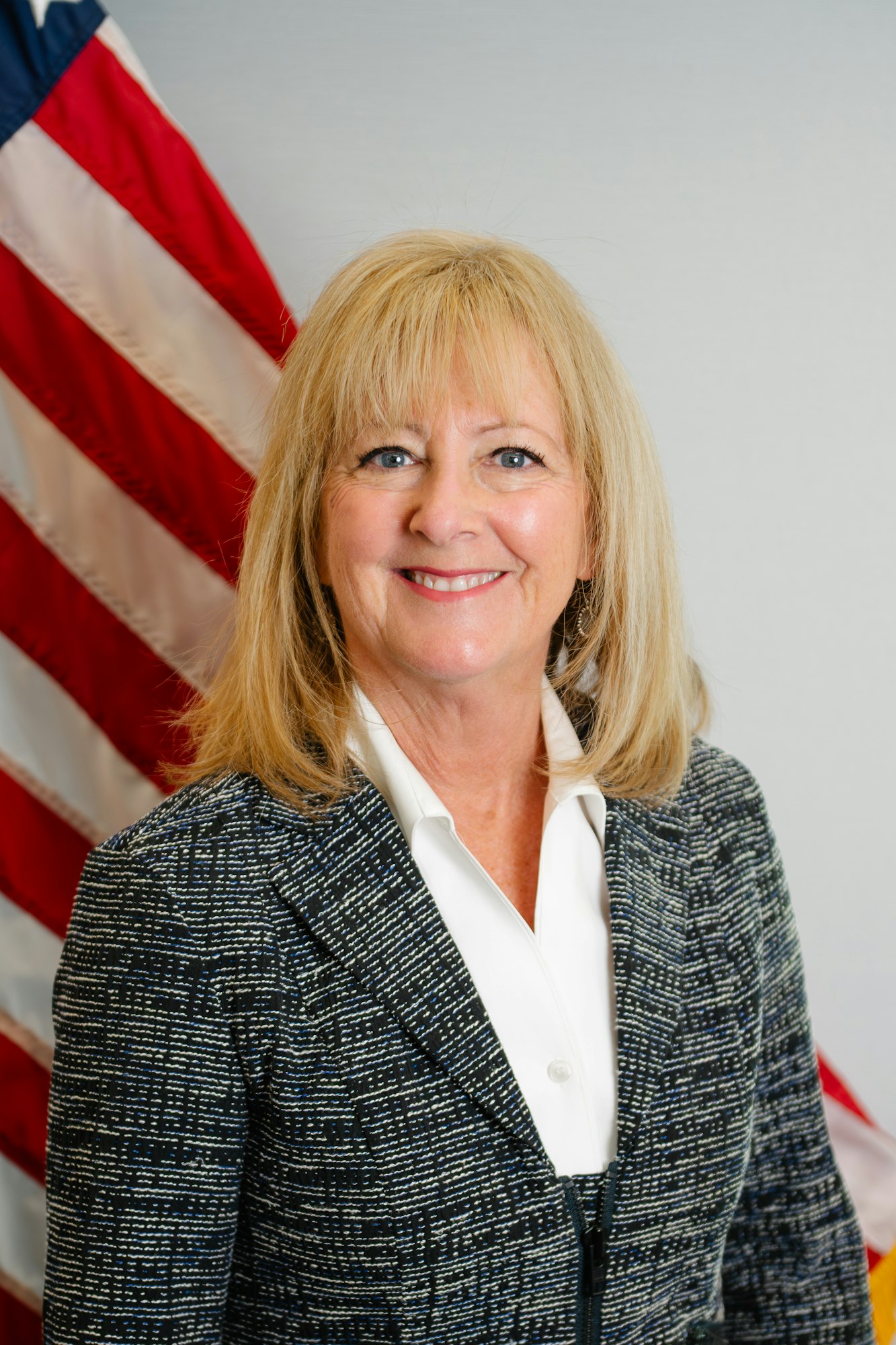 A smiling woman with blonde hair, wearing a patterned blazer, stands in front of an American flag.