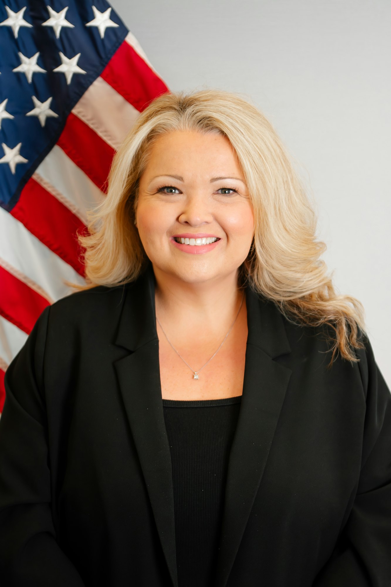 The image features a smiling woman in a black blazer, standing in front of an American flag backdrop.