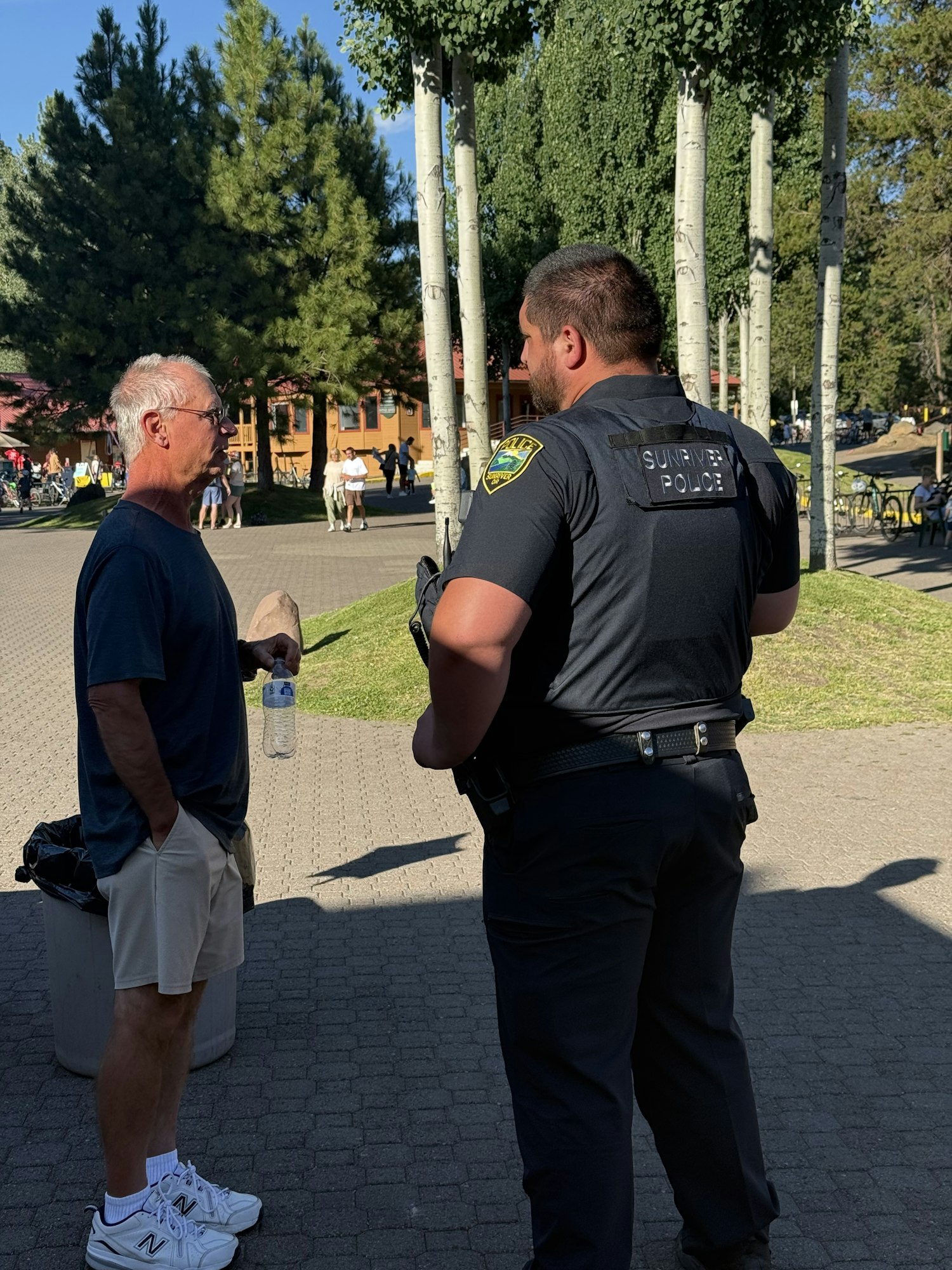 A police officer and a man converse on a pathway with trees in the background.