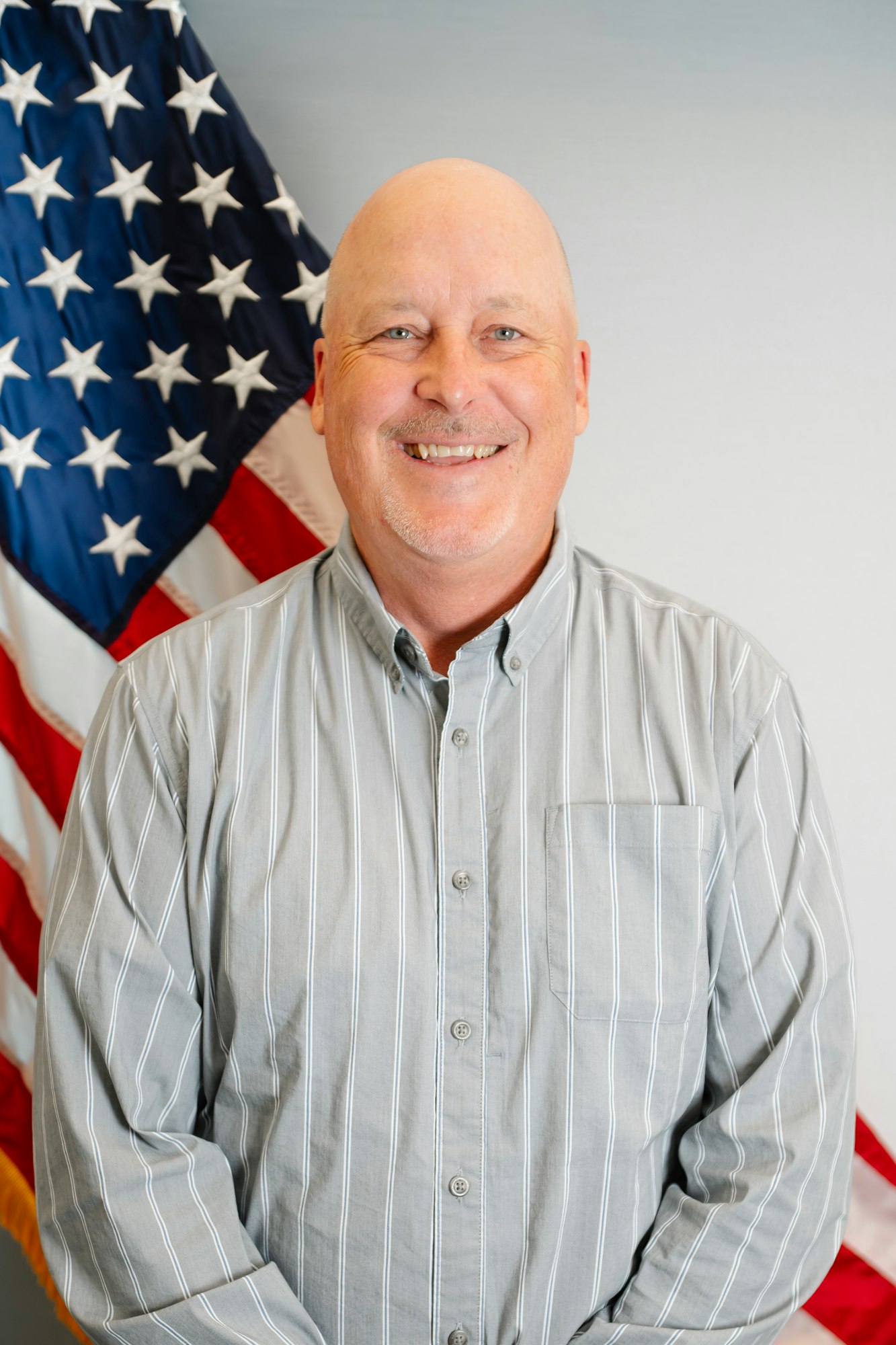 A smiling man in a striped gray shirt stands in front of an American flag backdrop.