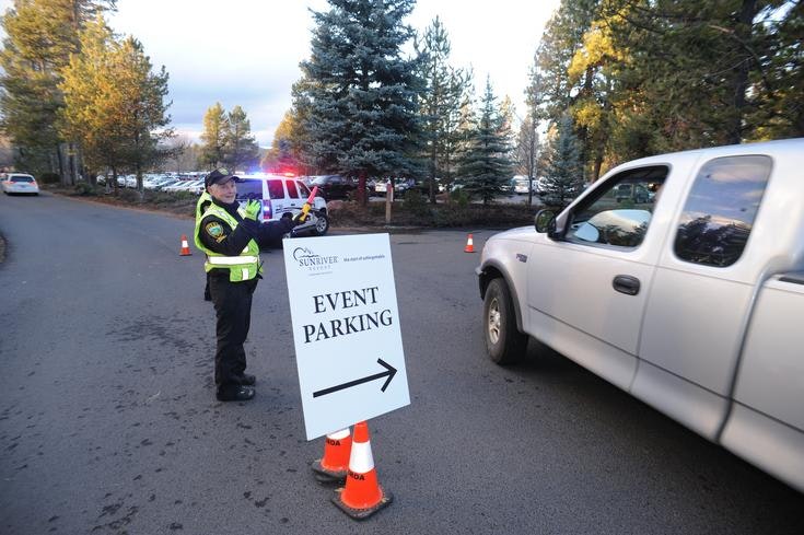 An officer directing traffic to event parking with a sign, vehicles, and police car lights in the background.