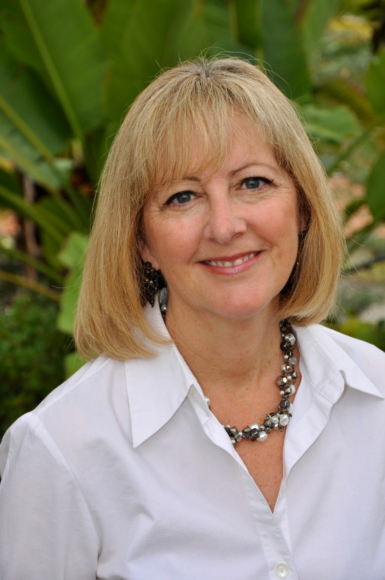 A person with short blonde hair, smiling, in a white shirt and necklace, with green leaves in the background.