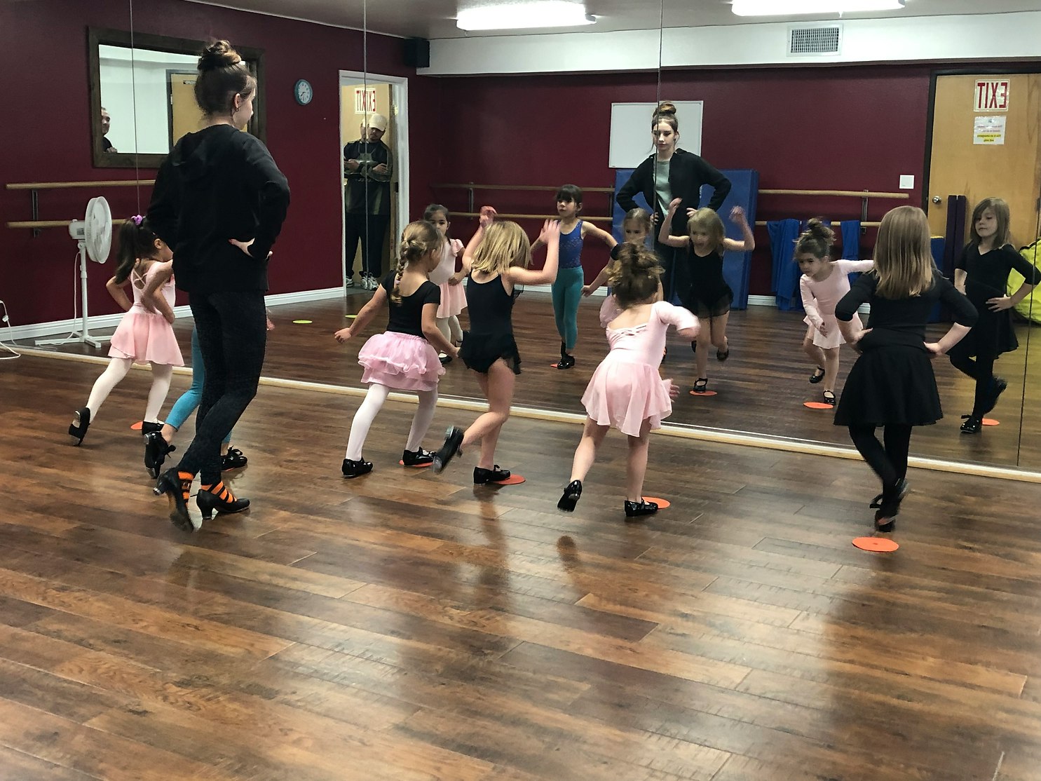Children practicing dance in a studio with mirrored walls, led by an instructor.
