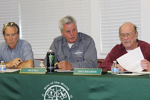 Three men seated at a table with a green tablecloth, discussing matters. One holds a document, and a can of drink is visible.