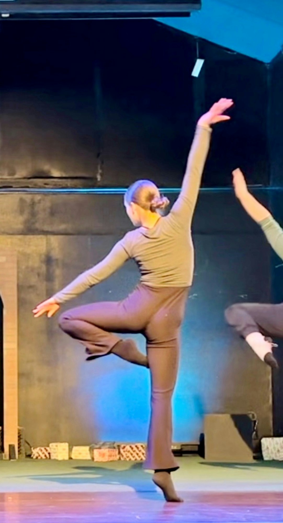 A dancer in a black outfit performing on stage, one leg raised, arm extended, against a dark background.