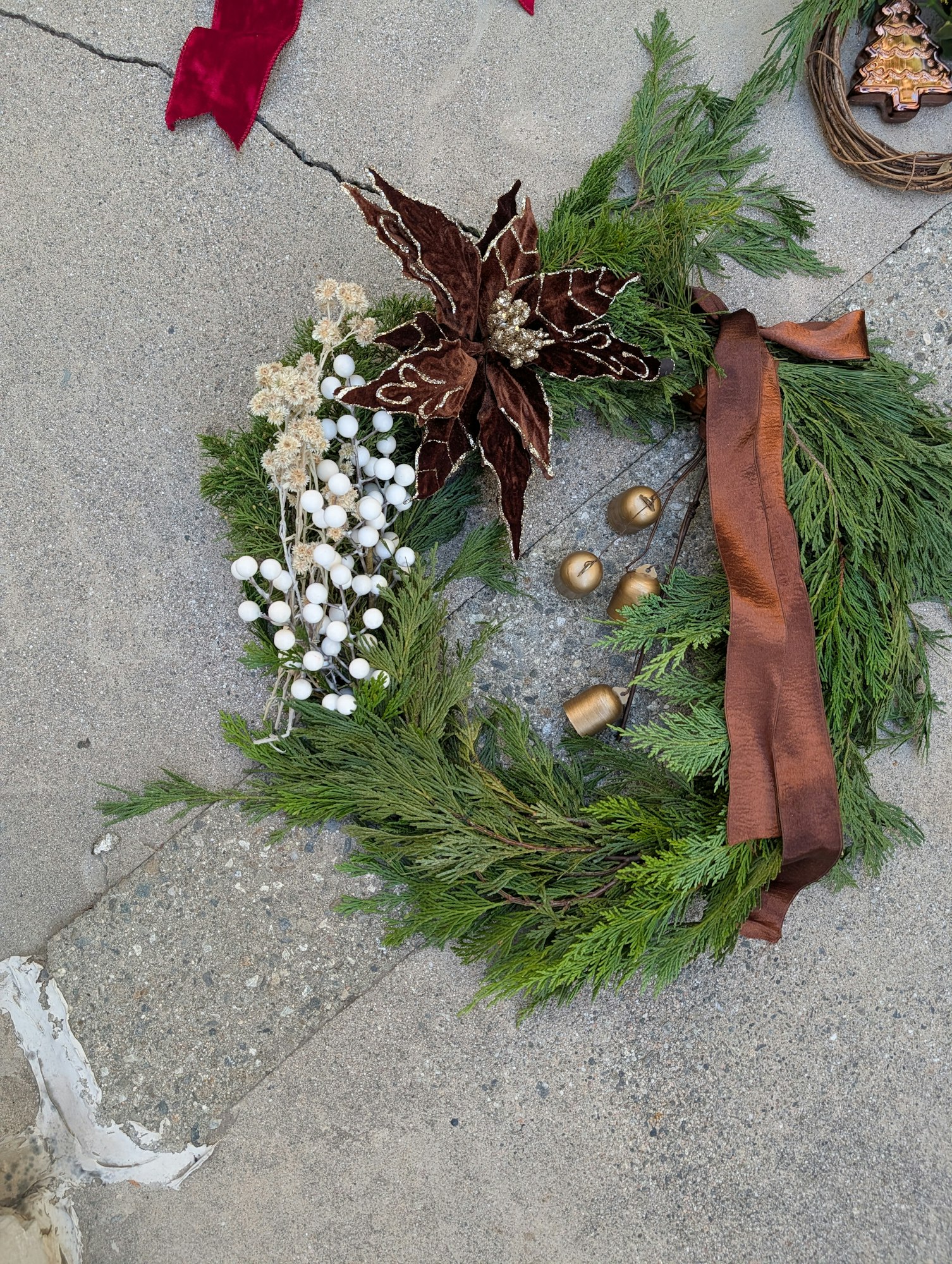 Festive wreath with greenery, a poinsettia, white berries, acorns, and a brown ribbon on a concrete surface.