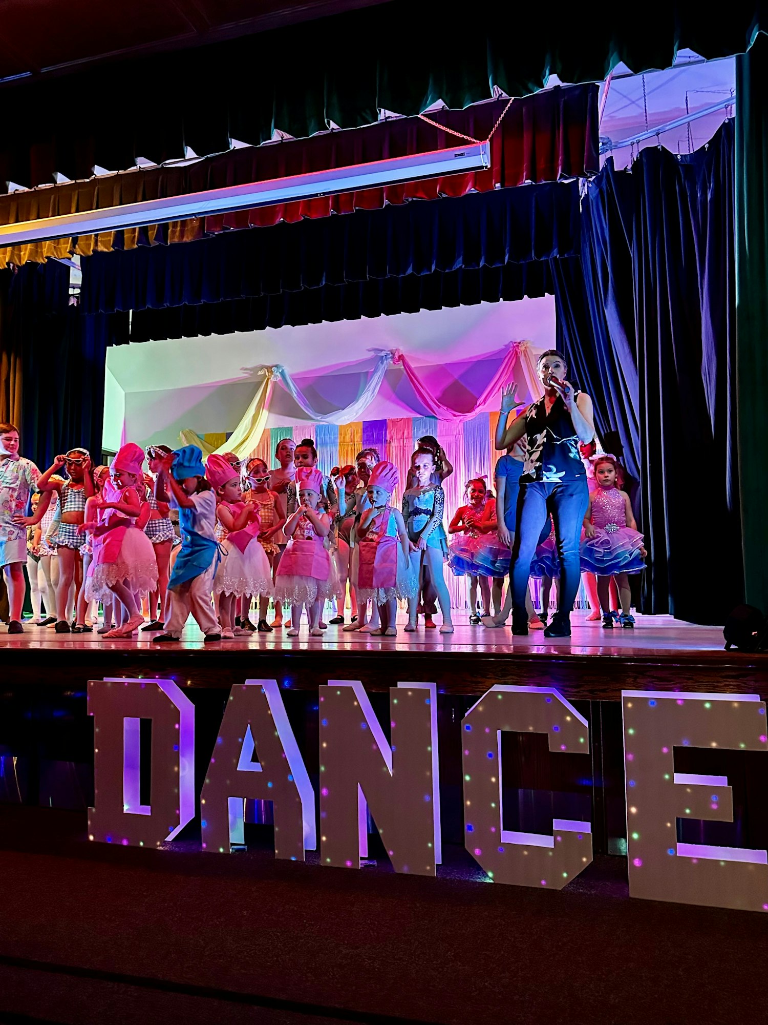 Children in colorful costumes on stage for a dance performance, with large "DANCE" letters lit up below.