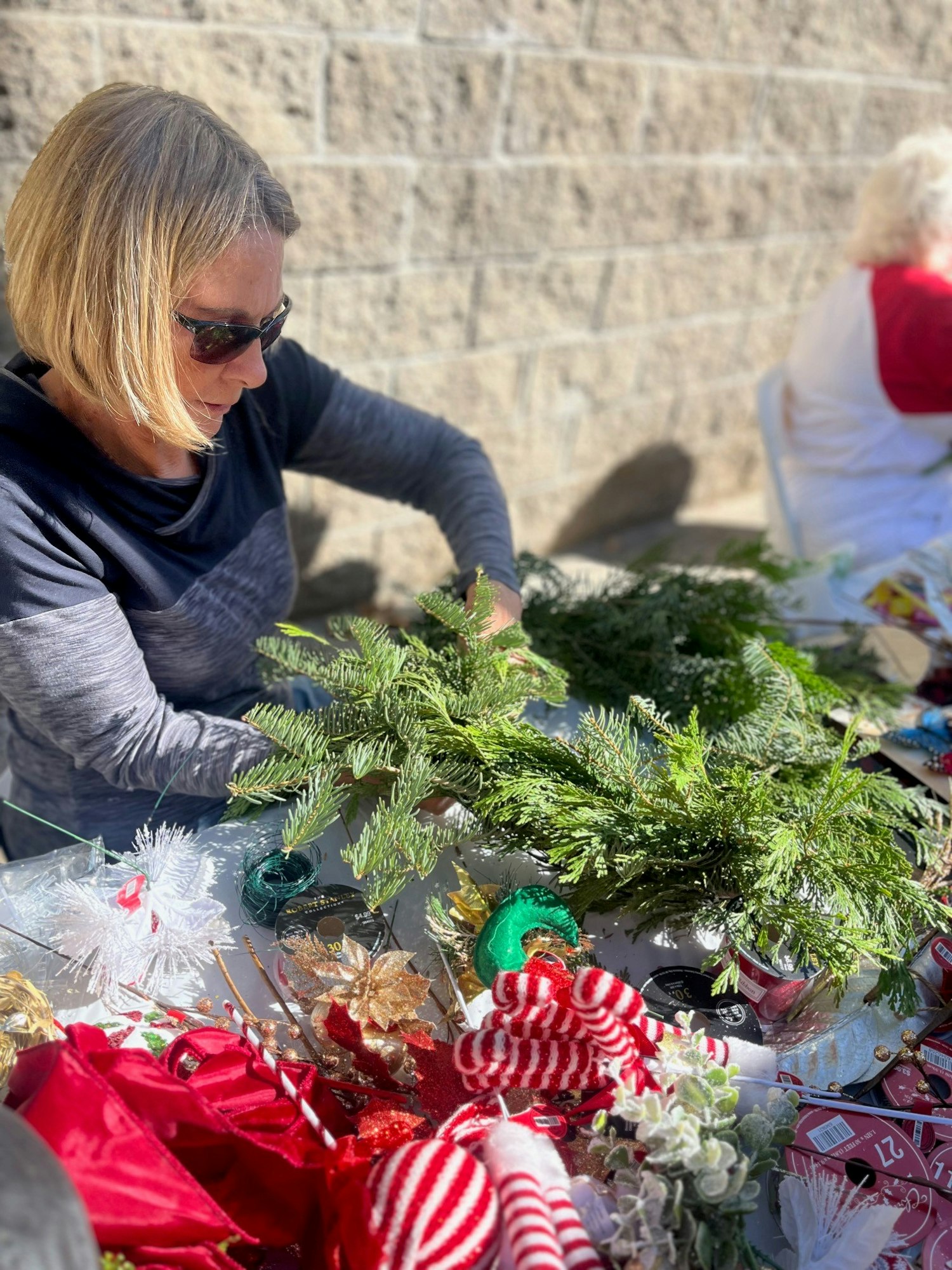 A woman wearing sunglasses arranges festive decorations and greenery at an outdoor table, with holiday-themed items around her.